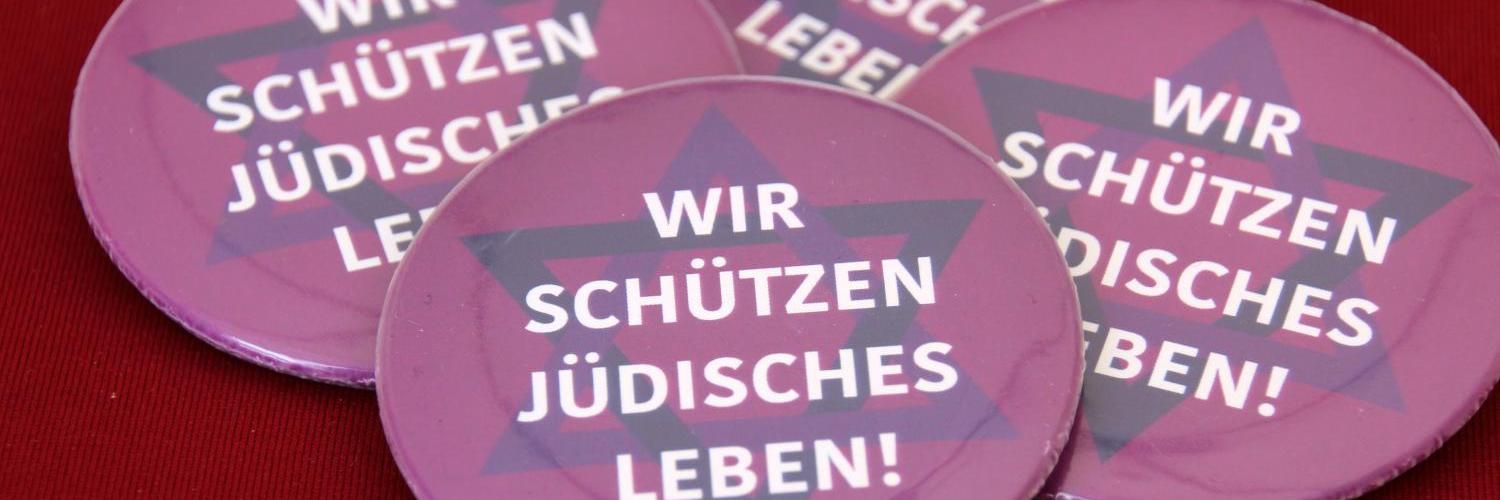 Vier lilafarbene Buttons mit der Aufschrift „Wir schützen jüdisches Leben!“ auf rotem Untergrund.