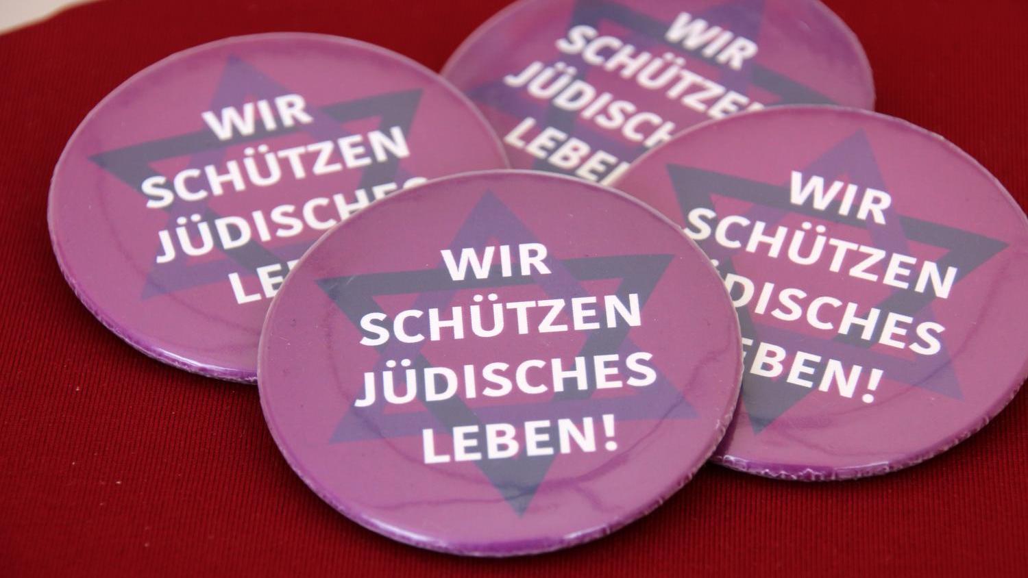 Vier lilafarbene Buttons mit der Aufschrift „Wir schützen jüdisches Leben!“ auf rotem Untergrund.