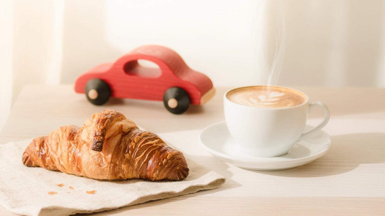 Tasse Kaffee, Croissant und rotes Spielzeugauto aus Holz auf einem Tisch