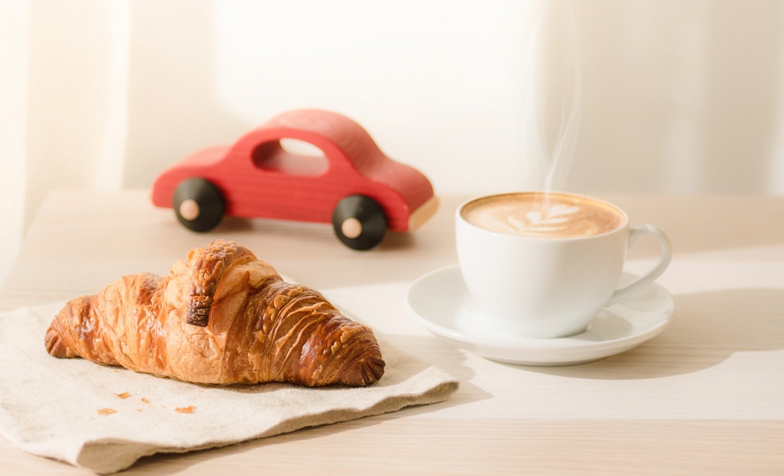 Tasse Kaffee, Croissant und rotes Spielzeugauto aus Holz auf einem Tisch