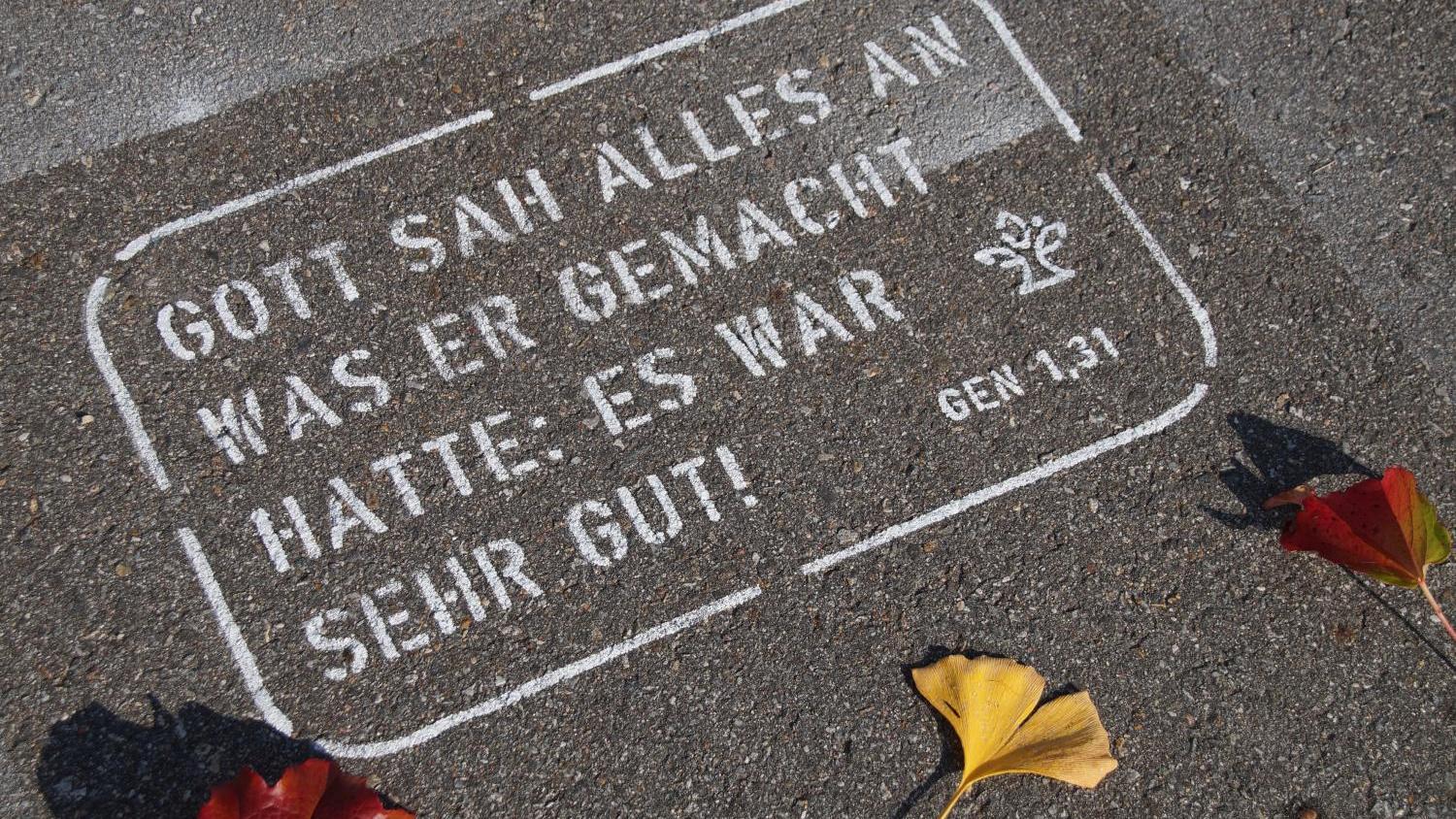 Weißer Bibelvers auf Asphalt, umgeben von bunten Herbstblättern.
