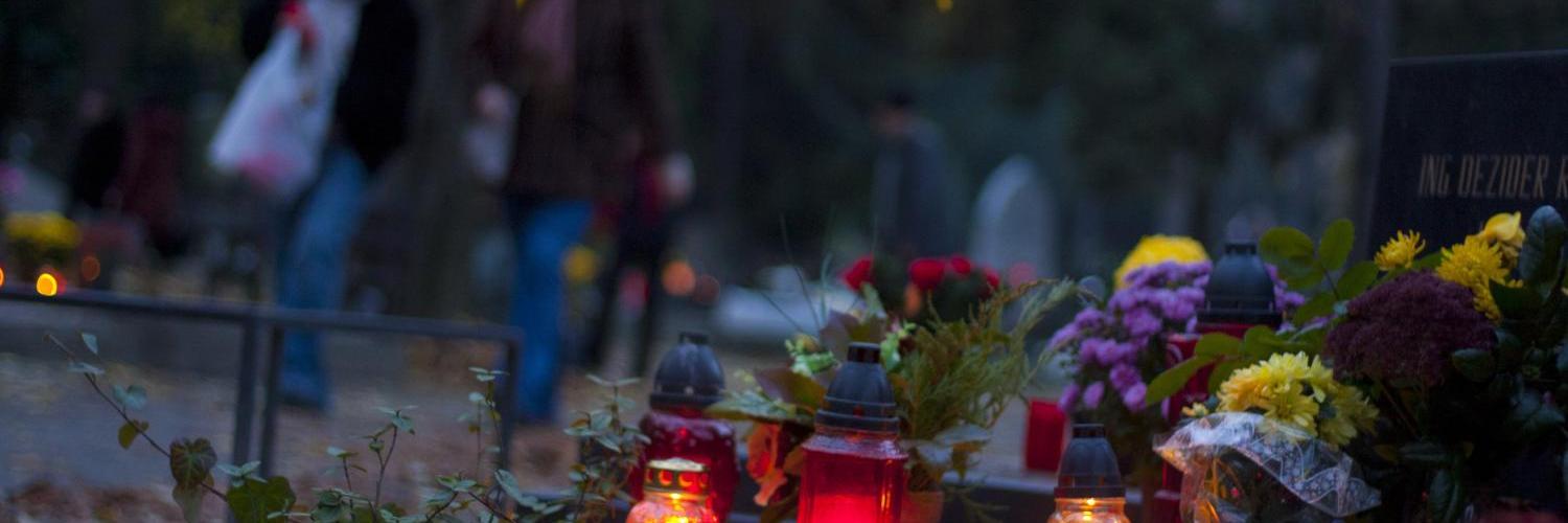 Kerzen und Blumen auf einem Grabstein, verschwommene Menschen im Hintergrund.
