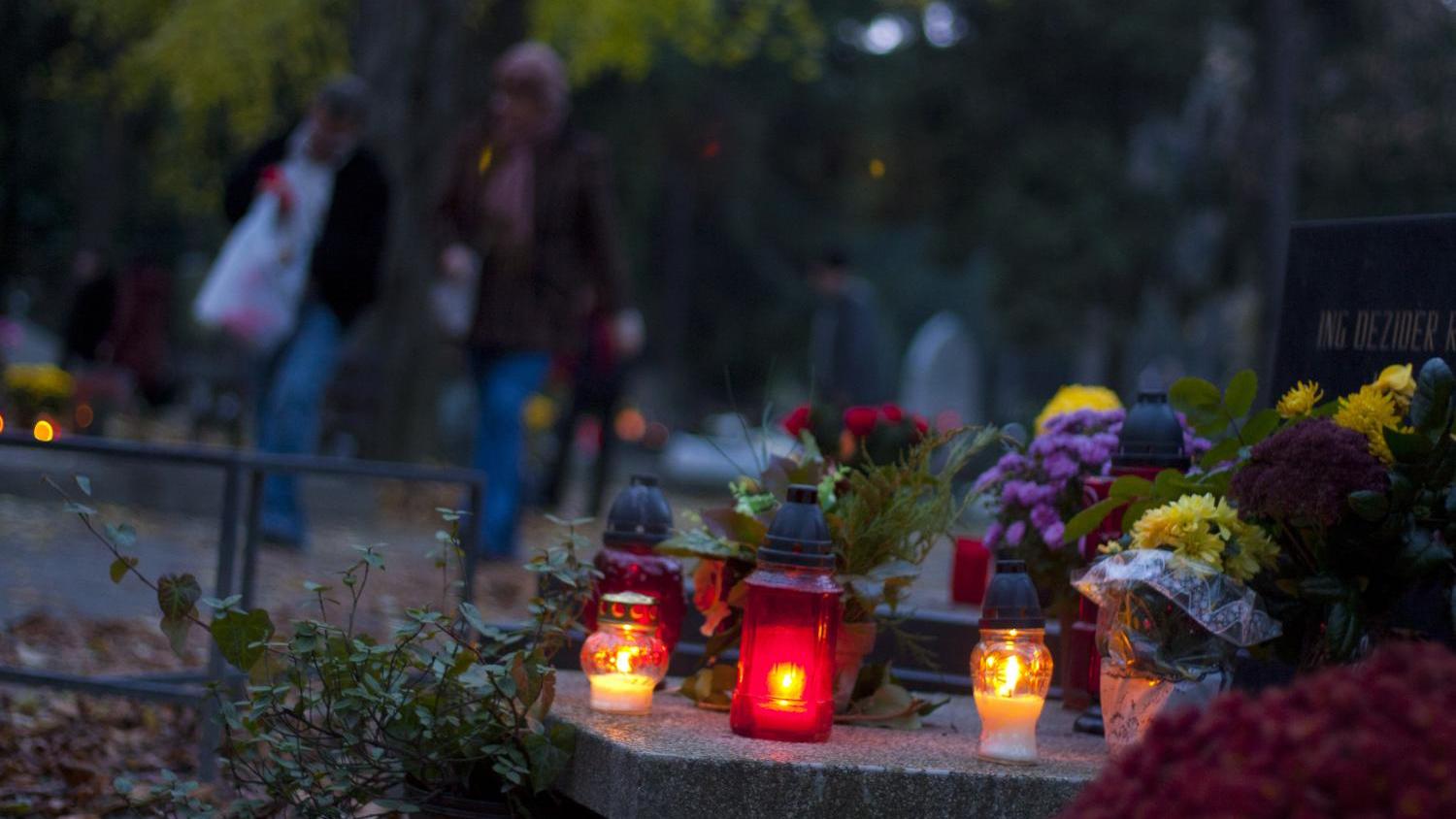Kerzen und Blumen auf einem Grabstein, verschwommene Menschen im Hintergrund.
