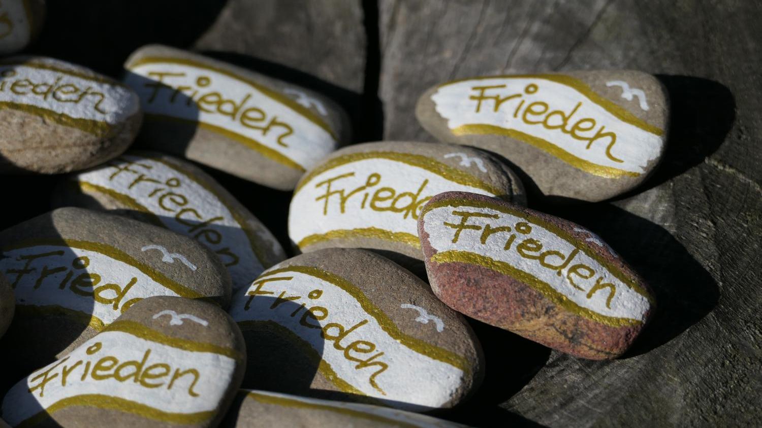 Bemalte Steine mit dem Wort „Frieden“ auf Holzuntergrund.