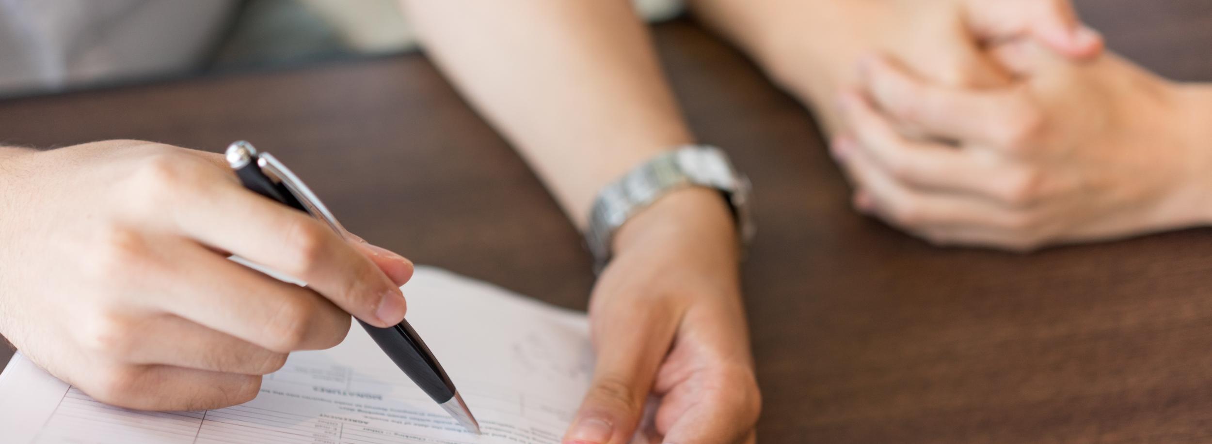 Man sieht Hände einer Frau und eines Mannes, Papiere. Der Mann hält einen Stift. Vermutlich wird ein Vertrag ausgehandelt.
