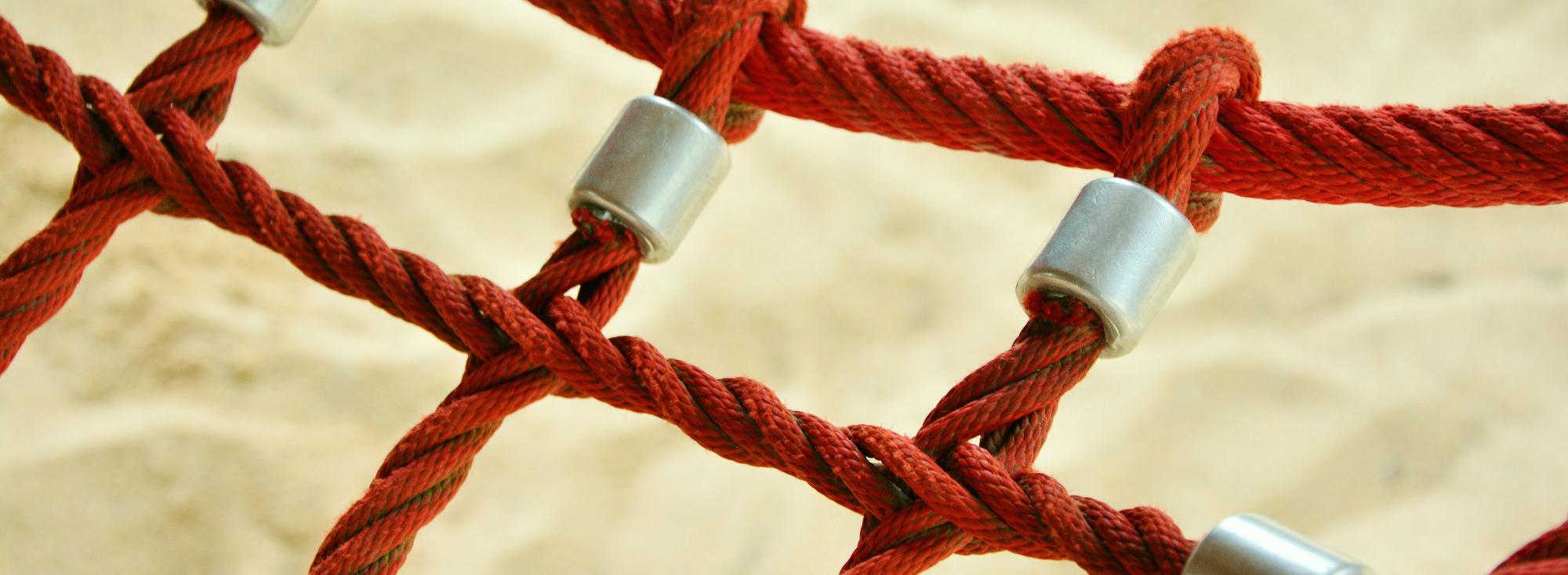 Rotes Kletterseil mit Metallverbindern vor sandigem Hintergrund