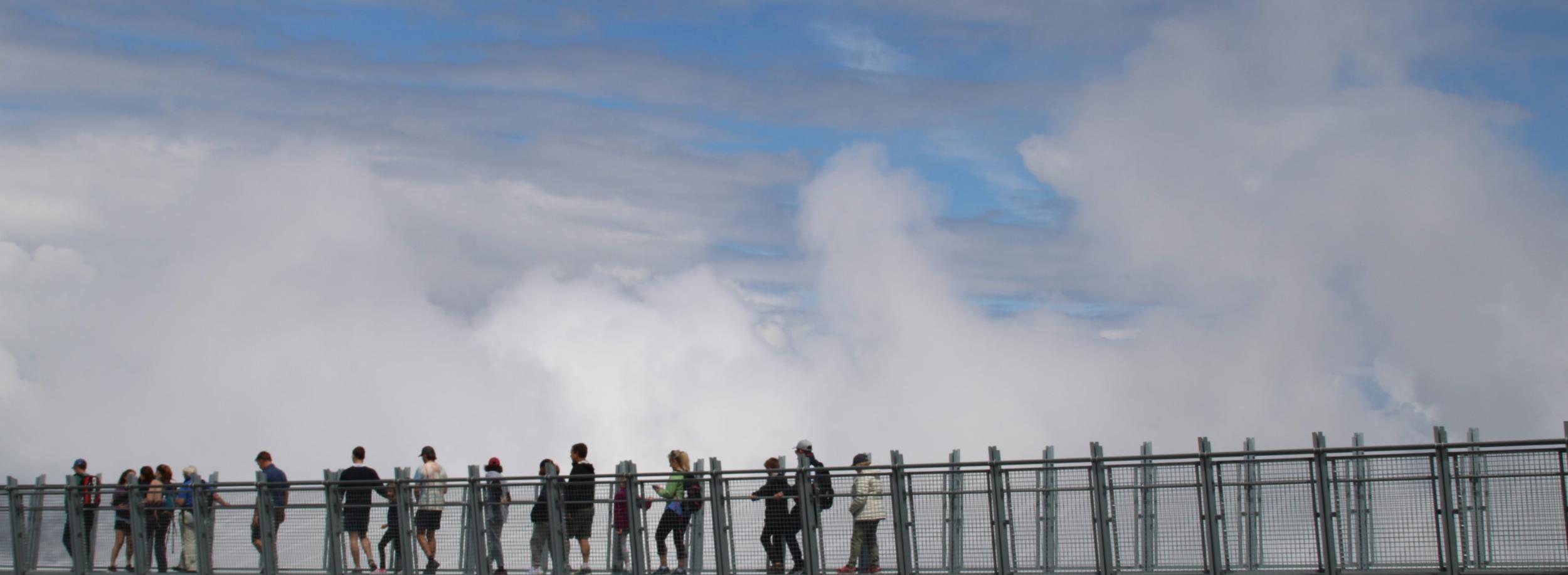 Menschen gehen über eine Hängebrücke. Im Hintergrund sind Wolken zu sehen
